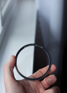 A hand is holding a circular camera lens filter with the markings 'B+W 72 010 UV-Haze 1x MRC' visible. The background is a blurred window with light shining through.