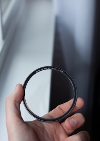 A hand is holding a circular camera lens filter with the markings 'B+W 72 010 UV-Haze 1x MRC' visible. The background is a blurred window with light shining through.
