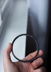 A hand is holding a circular camera lens filter with the markings 'B+W 72 010 UV-Haze 1x MRC' visible. The background is a blurred window with light shining through.