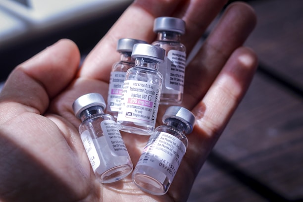 A hand holding several small glass vials with labels and silver caps. The vials contain a clear liquid and have medical information printed on the labels. The background is slightly blurred, suggesting an indoor setting with natural light.