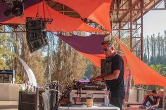 A DJ is performing on a stage set up outdoors, surrounded by vibrant orange and purple fabric structures. Large speakers are prominently displayed, and a person can be seen sitting on the right side of the image. The setting appears to be festive and lively, with trees and a clear sky in the background.