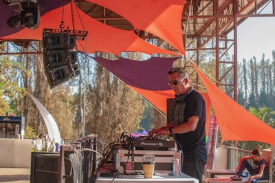 A DJ is performing on a stage set up outdoors, surrounded by vibrant orange and purple fabric structures. Large speakers are prominently displayed, and a person can be seen sitting on the right side of the image. The setting appears to be festive and lively, with trees and a clear sky in the background.