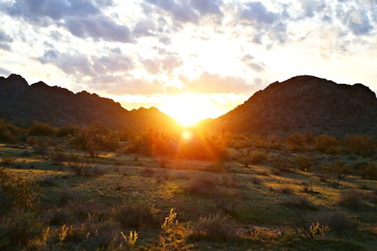 A vibrant sunrise peeks over the silhouette of a mountainous landscape, casting a warm golden glow over the desert terrain. Fluffy clouds scatter across the sky, reflecting the sunlight, while sparse desert vegetation adds texture to the foreground.