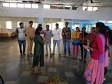 A group of people standing in a semi-circle in a large, open indoor space. The room has a tiled floor and high windows that let in natural light. Two individuals are in the foreground, one holding a microphone. The others are casually dressed and appear to be listening or engaged in a discussion.