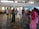 A group of people standing in a semi-circle in a large, open indoor space. The room has a tiled floor and high windows that let in natural light. Two individuals are in the foreground, one holding a microphone. The others are casually dressed and appear to be listening or engaged in a discussion.