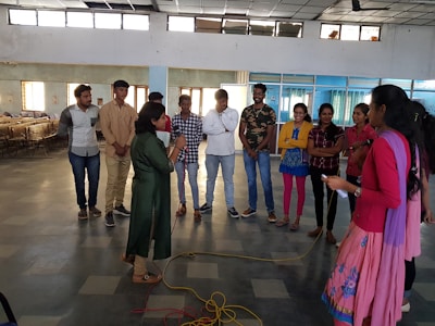 A group of people standing in a semi-circle in a large, open indoor space. The room has a tiled floor and high windows that let in natural light. Two individuals are in the foreground, one holding a microphone. The others are casually dressed and appear to be listening or engaged in a discussion.