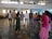 A group of people standing in a semi-circle in a large, open indoor space. The room has a tiled floor and high windows that let in natural light. Two individuals are in the foreground, one holding a microphone. The others are casually dressed and appear to be listening or engaged in a discussion.