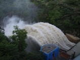 A powerful surge of water flows over a dam, creating a foamy and turbulent cascade. The surrounding area is lush with dense green foliage, and there is a structure with a blue facade in the foreground, suggesting a controlled water management facility.