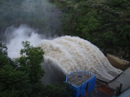 A powerful surge of water flows over a dam, creating a foamy and turbulent cascade. The surrounding area is lush with dense green foliage, and there is a structure with a blue facade in the foreground, suggesting a controlled water management facility.