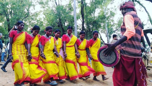 A group of women dressed in vibrant yellow and red traditional attire are performing a dance in a natural outdoor setting. They are holding hands and moving in unison, while a person wearing a red and orange outfit plays a drum nearby. Trees and other onlookers are visible in the background.