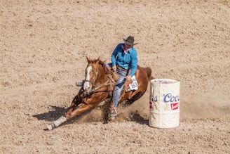 A cowboy is performing a barrel racing maneuver with a horse in a rodeo setting, kicking up dust as they make a sharp turn around a branded barrel.