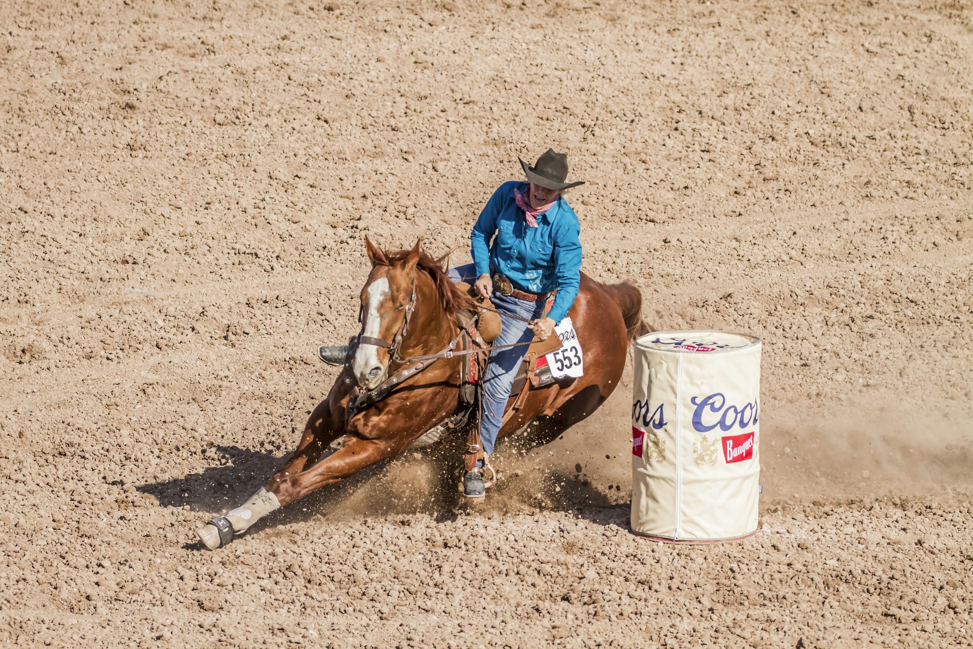 A cowboy is performing a barrel racing maneuver with a horse in a rodeo setting, kicking up dust as they make a sharp turn around a branded barrel.