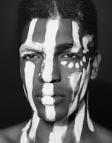 A black and white portrait features a person with an intricate pattern of white paint applied to the face and chest. The paint contrasts sharply against the skin, creating striking vertical stripes and shapes that give an artistic and tribal appearance.