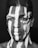 A black and white portrait features a person with an intricate pattern of white paint applied to the face and chest. The paint contrasts sharply against the skin, creating striking vertical stripes and shapes that give an artistic and tribal appearance.