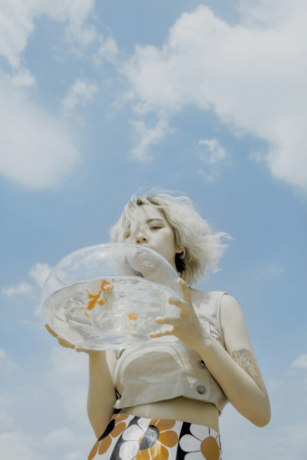 woman holding round clear glass fishbowl with live fish under white and blue sky