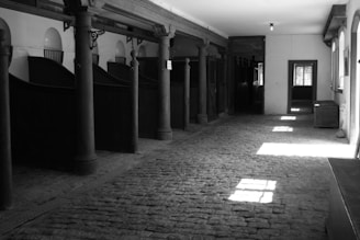 A black and white photograph depicts a stable or horse barn interior with empty stalls and cobblestone flooring. Sunlight filters through windows, casting rectangular light patches on the floor. Dark wooden posts and beams line the hallway, supporting the structure.