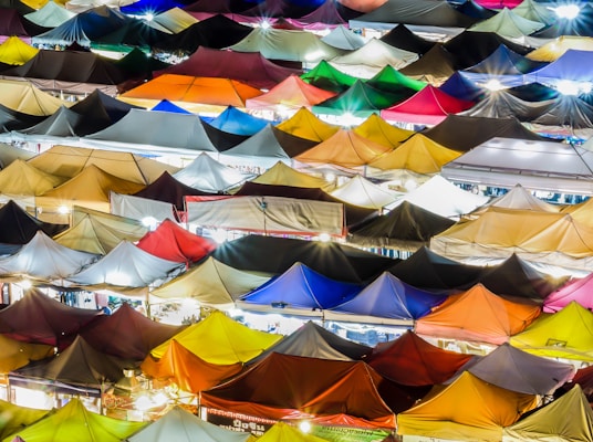 A vibrant array of colorful market tents densely packed together, illuminated by bright lights, creating a kaleidoscope of hues. The tents form a patchwork pattern across the image, suggesting a lively evening market.