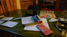 A table with various items including a book titled 'Istanbul' by Orhan Pamuk, a smartphone with a peach-themed case, scattered papers, a pen, and a cup with a saucer. The table has a rustic, wooden look, and the wall in the background is made of exposed brick, suggesting a cozy cafe atmosphere.