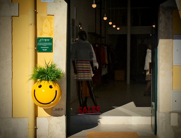 An entrance to a clothing store with a cheerful yellow smiley face decoration, which features spiky green foliage on top, resembling hair. The entrance displays a mannequin dressed in a skirt and sweater. The word 'SALES' is visible on the glass door. Pendant lights are hanging inside the store.