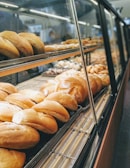A bakery display case filled with various types of bread, including rolls and loaves, arranged neatly on metal racks within a glass enclosure.