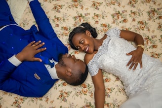 A couple lies on a floral-patterned surface, facing each other with smiles. The woman is wearing a white lace dress, while the man is dressed in a blue suit. Their hands are gently placed on each other's chests, conveying a sense of intimacy and happiness.