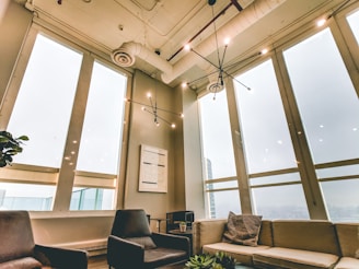 An indoor modern lounge area featuring large, floor-to-ceiling windows that allow natural light to flood the room. The space is furnished with comfortable chairs and a sofa arranged around a small table with a plant. The ceiling has exposed ducts and stylish, minimalistic light fixtures. A piece of abstract art hangs on one of the walls.