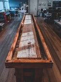 A long wooden shuffleboard table stretches through an office space with wood flooring. The room features modern office furniture, including desks with computers, chairs, and a kitchenette area in the background. Natural light pours in through large windows, casting shadows across the room and table.