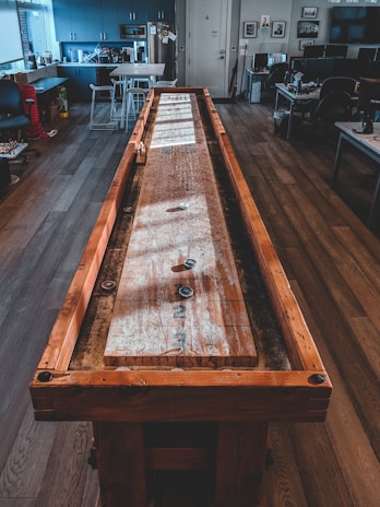A long wooden shuffleboard table stretches through an office space with wood flooring. The room features modern office furniture, including desks with computers, chairs, and a kitchenette area in the background. Natural light pours in through large windows, casting shadows across the room and table.