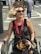A person in a wheelchair is dressed in stylish and colorful attire with a sequined black top, vibrant patterned pants, and bold accessories including a large red necklace and bejeweled sunglasses. Their hair is styled in an elaborate way, adorned with a decorative headband featuring feathers. They are outdoors on a city street, with pedestrians and a crosswalk visible in the background.