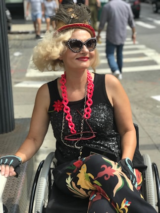 A person in a wheelchair is dressed in stylish and colorful attire with a sequined black top, vibrant patterned pants, and bold accessories including a large red necklace and bejeweled sunglasses. Their hair is styled in an elaborate way, adorned with a decorative headband featuring feathers. They are outdoors on a city street, with pedestrians and a crosswalk visible in the background.