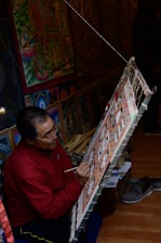 An artist is seated on the floor, focused intently on painting a detailed, colorful artwork displayed on a slanted easel. The room is filled with vibrant, intricate art pieces, suggesting a workshop or studio. The surroundings include more paintings and art supplies, indicating a creative and dedicated atmosphere.
