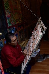 An artist is seated on the floor, focused intently on painting a detailed, colorful artwork displayed on a slanted easel. The room is filled with vibrant, intricate art pieces, suggesting a workshop or studio. The surroundings include more paintings and art supplies, indicating a creative and dedicated atmosphere.
