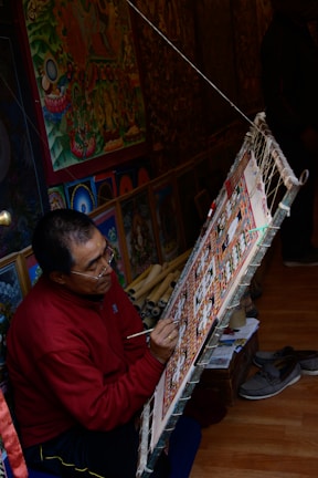 An artist is seated on the floor, focused intently on painting a detailed, colorful artwork displayed on a slanted easel. The room is filled with vibrant, intricate art pieces, suggesting a workshop or studio. The surroundings include more paintings and art supplies, indicating a creative and dedicated atmosphere.