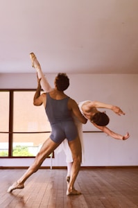 Two ballet dancers are performing an elegant pose in a dance studio. The male dancer is supporting the female dancer as she leans backward gracefully with one leg extended high. The room has a large window that lets in natural light.