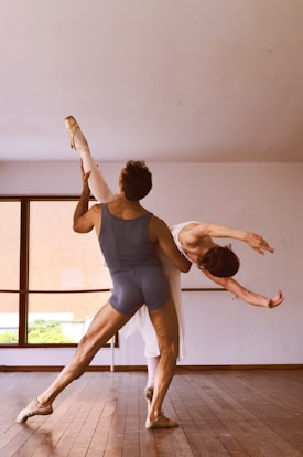 Two ballet dancers are performing an elegant pose in a dance studio. The male dancer is supporting the female dancer as she leans backward gracefully with one leg extended high. The room has a large window that lets in natural light.