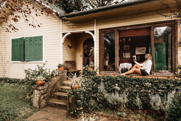 A woman is sitting on the windowsill of a charming, rustic house with yellow wooden siding and green shutters. The window is large and open, displaying an inviting interior room. She is relaxed, reading a magazine, with her legs crossed. The house is surrounded by lush greenery, including potted plants and vines creeping up the wall. Stone steps lead up to the front door, enhancing the quaint and peaceful setting.