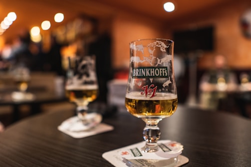 A close-up of a beer glass with 'Brinkhoff's No.1' insignia sits on a table in a dimly lit bar. The background is softly blurred, showing a warm, inviting atmosphere with out-of-focus lights and a relaxed setting.