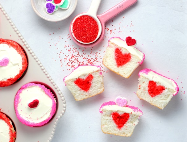 Cupcakes decorated with red and pink heart-shaped toppers and designs. The cupcakes are cut to reveal a red heart shape inside. Surrounding them are bowls of sprinkles and heart-shaped decorations.
