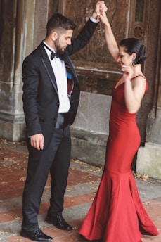 A couple engaged in a dance, with the man wearing a formal black suit and bow tie and the woman in a long red dress. They are standing on a tiled floor with dry leaves scattered around, possibly in an outdoor or historical setting with stone walls.