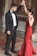 A couple engaged in a dance, with the man wearing a formal black suit and bow tie and the woman in a long red dress. They are standing on a tiled floor with dry leaves scattered around, possibly in an outdoor or historical setting with stone walls.