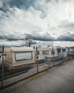 A row of RVs or caravans is parked behind a wire fence. The overcast sky is filled with large, fluffy clouds. A lone tree without leaves stands next to the caravans, casting a stark silhouette.