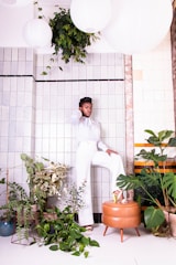 A stylish individual stands confidently against a tiled wall, surrounded by lush green plants. They are dressed in a sophisticated white outfit and posed with one leg resting on a brown leather ottoman. Large white paper lanterns hang from above, adding to the modern and elegant setting.