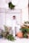 A stylish individual stands confidently against a tiled wall, surrounded by lush green plants. They are dressed in a sophisticated white outfit and posed with one leg resting on a brown leather ottoman. Large white paper lanterns hang from above, adding to the modern and elegant setting.