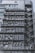 Numerous air conditioning units are densely packed on the exterior wall of a multi-story building. The image shows a very urban setting, with a grid-like arrangement of metal platforms supporting the units. Adjacent to this setup is a zigzagging metal staircase, leading to various floors of the building. The overall appearance is industrial and cluttered.