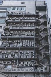 Numerous air conditioning units are densely packed on the exterior wall of a multi-story building. The image shows a very urban setting, with a grid-like arrangement of metal platforms supporting the units. Adjacent to this setup is a zigzagging metal staircase, leading to various floors of the building. The overall appearance is industrial and cluttered.