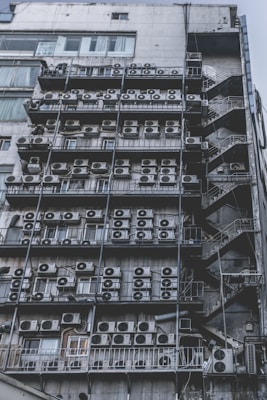 Numerous air conditioning units are densely packed on the exterior wall of a multi-story building. The image shows a very urban setting, with a grid-like arrangement of metal platforms supporting the units. Adjacent to this setup is a zigzagging metal staircase, leading to various floors of the building. The overall appearance is industrial and cluttered.