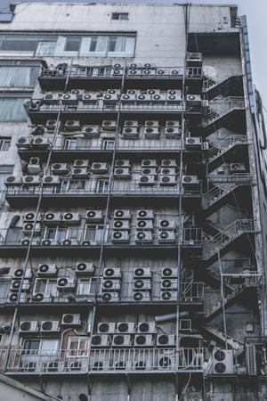 Numerous air conditioning units are densely packed on the exterior wall of a multi-story building. The image shows a very urban setting, with a grid-like arrangement of metal platforms supporting the units. Adjacent to this setup is a zigzagging metal staircase, leading to various floors of the building. The overall appearance is industrial and cluttered.