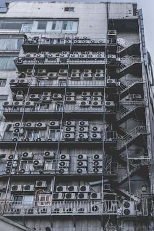 Numerous air conditioning units are densely packed on the exterior wall of a multi-story building. The image shows a very urban setting, with a grid-like arrangement of metal platforms supporting the units. Adjacent to this setup is a zigzagging metal staircase, leading to various floors of the building. The overall appearance is industrial and cluttered.
