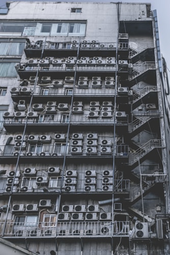 Numerous air conditioning units are densely packed on the exterior wall of a multi-story building. The image shows a very urban setting, with a grid-like arrangement of metal platforms supporting the units. Adjacent to this setup is a zigzagging metal staircase, leading to various floors of the building. The overall appearance is industrial and cluttered.