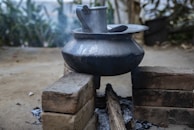 A metal pot sits atop a simple outdoor cooking setup, constructed using bricks and fueled by burning wood. Ashes and charred wood lie underneath the pot, while smoke rises slightly from the makeshift stove. The background is blurred and features greenery, suggesting a rural setting.